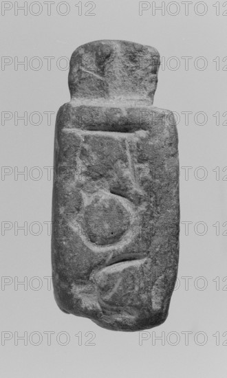 Christian amulet with Greek inscription. Limestone amulet in the shape of an elongated hexagon with Greek text on the sides and bottom. Upper edge has a protrusion with a drilled hole for suspension. The letters on the five edges form a coherent Greek inscription with magical meaning. 2nd–3rd century, found in Miletus. Amulet; height 5.2 cm, width 2.3 cm., A vertically oriented stone object with a rough, weathered surface is centered against a plain background; the stone has an irregular rectangular shape with a rounded top section and a wider lower body, showing carved depressions and raised areas that form circular and curved shapes on its face; the surface texture appears pitted and grainy with shadows in the carved areas and smoother worn patches on the edges; visible colors include shades of gray and muted black tones across the stone and a pale gray background.