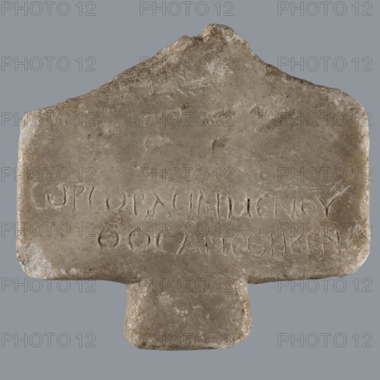 Greek dedicatory inscription, unknown author, Horus myth., tombstone inscription, Late Roman Empire period, Egypt, Greek inscription; height 12.1 cm, width 13.4 cm, depth 1.2 cm, A roughly rectangular stone slab with a protruding tab at its bottom and an irregular peak at its top, shown against a plain gray background. The stone surface is weathered and textured with worn edges and small pits, displaying a palette of beige, light brown, gray, and hints of off-white. There are shallow incised marks arranged in horizontal groupings on the lower half of the slab. The overall shape features softened corners and an uneven top contour, and the surface shows subtle variations in tone and slight areas of discoloration.