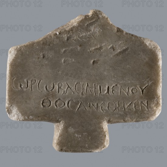 Greek dedicatory inscription; Horus myth.; tombstone inscription; Late Roman Empire period, Egypt; Greek inscription; height 12.1 cm, width 13.4 cm, depth 1.2 cm;, A roughly rectangular stone object with a protruding tab at the bottom, displayed against a gray background, showing an uneven, worn surface with chipped and rounded edges; the stone has a mostly beige and light brown tone with areas of darker brown and faint gray, and there are shallow carved grooves and indentations on the upper surface and a row of incised markings across the middle that appear recessed into the stone; the top edge is irregular and slightly peaked at the center, and the texture shows small pits and smoother areas suggesting surface wear.