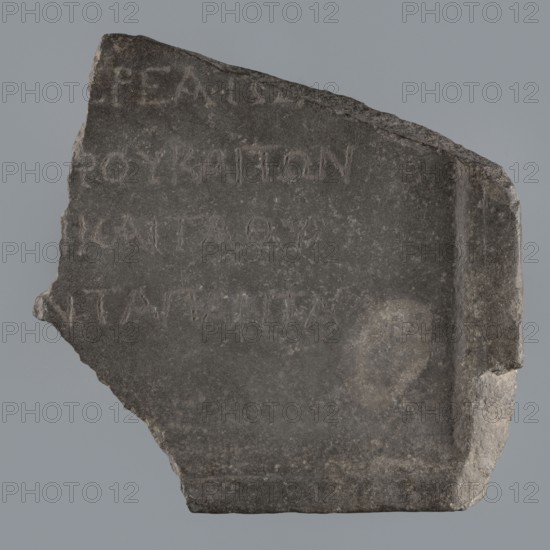 Fragment of a Greek dedicatory inscription, tombstone inscription, 70 BC–1 end of the Ptolemaic period or beginning of the Roman period, Egypt; Greek inscription; height 24.2 cm, width 24 cm, depth 4.7 cm., A roughly triangular fragment of dark gray stone with a rough, uneven edge on one side and a more rectangular, slightly beveled edge on the opposite side; the stone surface shows weathered texture with lighter gray patches and subtle raised areas where shallow incised marks are visible but not described; overall colors are dark gray, medium gray, and lighter gray with small areas of off-white at the chipped edges.