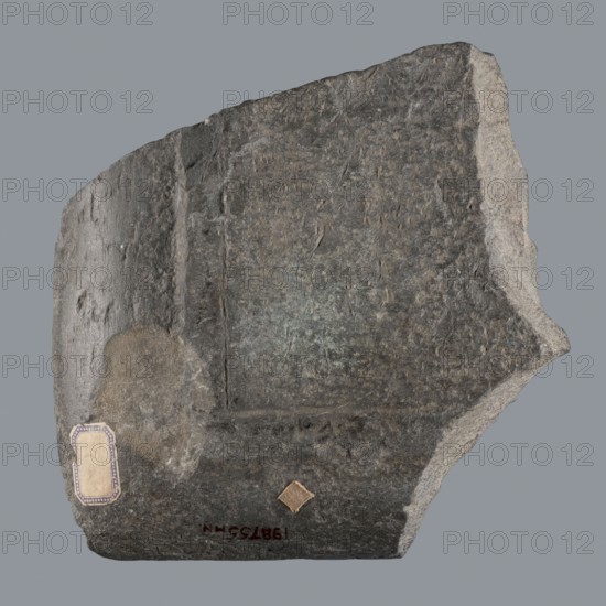 Fragment of a Greek dedicatory inscription, unknown author, tombstone inscription, 70 BC–1 end of the Ptolemaic period or beginning of the Roman period, Egypt, Greek inscription; height 24.2 cm, width 24 cm, depth 4.7 cm, A dark gray stone fragment with rough, uneven edges sits against a plain light gray background; the stone surface shows textured, mottled areas in shades of dark gray, medium gray, and lighter gray with a subtle greenish patina in places, and a rounded smoother patch of lighter gray on one side; there is a small beige and tan rectangular label with a patterned border and a small diamond-shaped tan patch adhered to the stone.