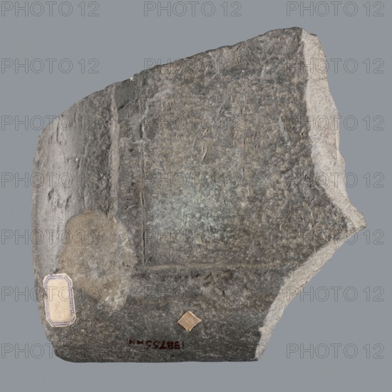 Fragment of a Greek dedicatory inscription, unknown creator/author, tombstone inscription, 70 BC–1 end of the Ptolemaic period or beginning of the Roman period, Egypt, Greek inscription, height 24.2 cm, width 24 cm, depth 4.7 cm, A roughly rectangular fragment of stone with irregular broken edges fills the frame, showing a textured surface with worn, carved areas and a recessed rectangular panel. The stone displays shades of gray, dark gray, light gray, and hints of muted brown, with a small patch of beige on one side and a small square patch of tan. The surface has rough, pitted areas, smoother worn sections, and visible striations and chips along the edges. The background is a uniform medium gray.