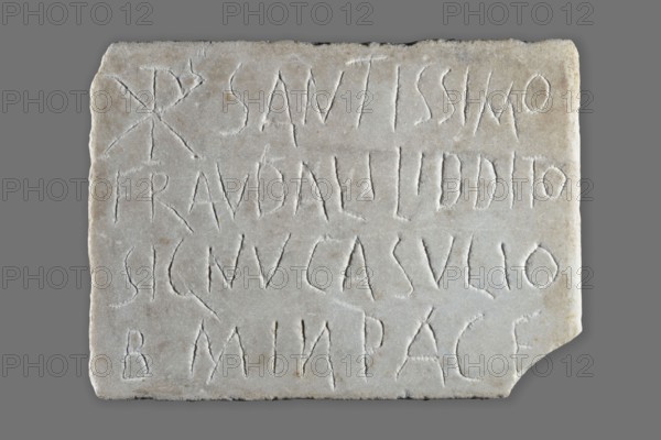Tombstone with Latin Christian inscription, unknown Roman workshop, 4th–5th century, Roman Empire 27 BC–476, slab; height 26 cm, width 19 cm, depth 2 cm, weight 2.65 kg, A rectangular pale stone slab with rough edges set against a solid gray background, showing shallow carved lines forming linear shapes across its surface; the stone is mostly off-white with areas of light beige and subtle gray veining, and there are a few small darker spots and slight surface weathering.