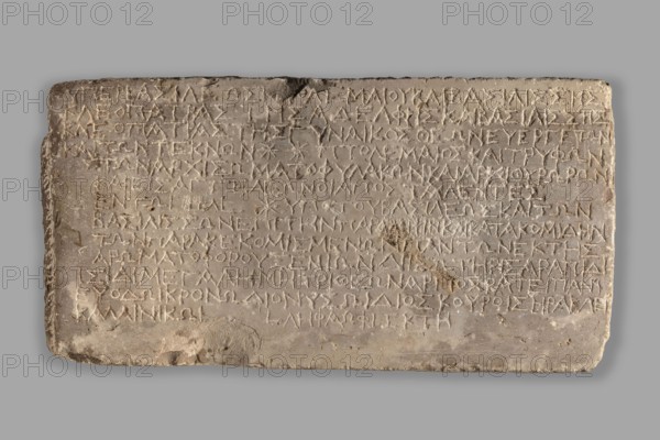 Greek dedicatory inscription, unknown author, Greek script; Ptolemy VIII Euergetes II Physkos ruler of Egypt; 170–116 BC; Greek inscriptions dedicating for many gods during the reign of Ptolemy VIII Euergetes II and his two wives and their children; issued by Ptolemy and Trypho, 30/10/133 BC; Coptos place of origin; inscription; height 25 cm, width 48 cm, depth 7 cm, weight 19.77 kg, A rectangular stone slab with a rough, weathered surface resting against a neutral gray background, showing a palette of pale beige, light gray, and warmer tan tones; the slab has worn, chipped edges and a few small darker brown spots, with shallow grooves and incisions across its face forming a dense pattern; the top edge is slightly irregular and the surface shows subtle variations in texture and color, including areas of smoother pale gray and rougher beige patches, as well as a few faint shadowed depressions and a softer gray cast along the lower edge.