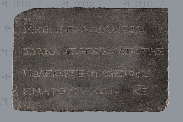 Greek dedicatory inscription, tombstone inscription, 2nd half of the 2nd century BC, Egypt; Greek inscription; height 22.0 cm, width 32.5 cm, depth 7.0 cm;, A rectangular dark gray stone slab with rough irregular edges sits against a lighter gray background, its surface showing a slightly uneven texture and small worn spots; a few shallow linear incisions are visible across the slab arranged in several horizontal rows, each incision appearing lighter in color than the surrounding stone and forming repeated angular shapes; the slab's top left and bottom right corners are slightly chipped, and the overall palette includes dark gray, light gray, and subtle brownish-gray variations.