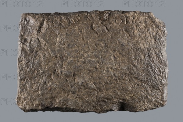 Greek dedicatory inscription, unknown author, tombstone inscription, 2nd half of the 2nd century BC, Egypt, Greek inscription; height 22.0 cm, width 32.5 cm, depth 7.0 cm, A roughly rectangular slab with a rough, uneven surface and irregular edges sits against a flat background; the slab displays a textured, pitted appearance with visible ridges and small depressions, and its surface shows mottled shades of brown and gray with hints of darker brown and occasional lighter beige areas; the edges are jagged and slightly chipped, and the overall shape is somewhat flat but with a rugged, natural contour.