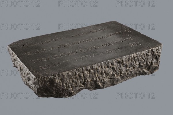 Greek dedicatory inscription, tombstone inscription, 2nd half of the 2nd century BC, Egypt; Greek inscription; height 22.0 cm, width 32.5 cm, depth 7.0 cm;, A flat, rectangular slab of dark stone with a rough, chiseled underside and textured edges rests against a plain gray background; the top surface is smoother and slightly uneven with a series of shallow incisions forming linear marks, and the stone shows colors including dark gray, charcoal, lighter gray highlights, and some brownish tones in the rougher areas.