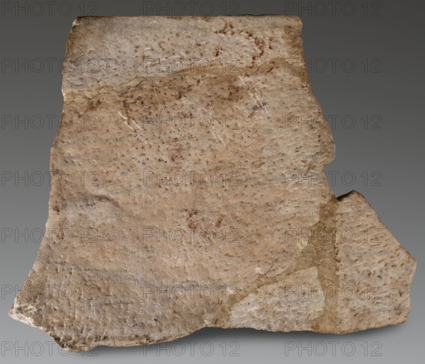 Fragment of a sarcophagus depicting a married couple, unknown Roman workshop; relief of marriage iconography; sarcophagus Roman, dated 200–220 formerly dated to the 230s of the 3rd century AD; Roman Empire 27 BC–476; materials and form: sarcophagus, height 73 cm, width 92 cm, depth 12 cm, weight 92 kg., A roughly triangular fragment of stone with an uneven, weathered surface occupies the image, showing a palette of beige, tan, light brown, and hints of gray with small darker brown speckles scattered across. The edges are irregular and chipped, with a visible break and a slightly protruding corner on one side where the interior texture appears coarser and more yellowish. The stone surface has subtle variations in tone and faint linear impressions, as well as areas that look smoother and slightly lighter near the top and bottom, and a few small spots of darker discoloration. The background is a smooth gradient of medium gray.