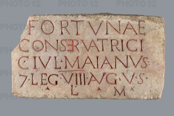 Slab with a Latin votive inscription for Fortuna Conservatrix, Caius Iulius Maianus 2nd–3rd century commissioner, Fortuna mythology, Latin inscriptions, baths, centurions, Germania Superior province, Roman legions. Limestone slab with a smoothed front surface and sides and an irregular back; upper left corner missing. Front surface carved with a 5-line inscription; letters were at one point painted red two paints used: a darker in most letters and a lighter, very thick paint in the letter E in line 2. Dated 130–259/260. Slab, votive inscription; width 33 cm, height 18 cm, thickness 6 cm., A rectangular stone slab with a rough, slightly chipped edge and a weathered surface sits against a plain gray background; the stone is pale beige with areas of darker beige and faint brownish spots, and it has carved grooves filled with a muted reddish pigment that form linear shapes across the surface; the carving appears arranged in several horizontal bands, with some letters showing wear and shall...