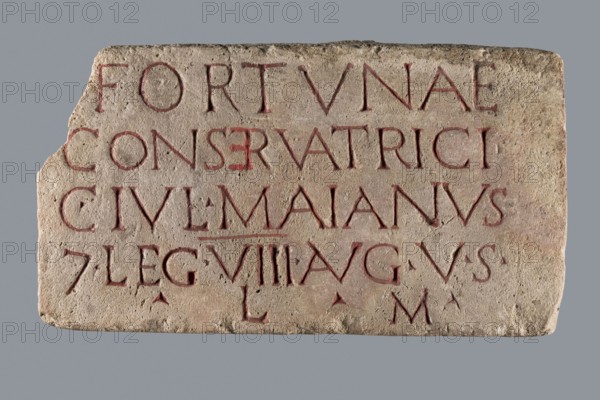 Slab with a Latin votive inscription for Fortuna Conservatrix, Caius Iulius Maianus 2nd–3rd century commissioner, Fortuna mythology, Latin inscriptions, baths, centurions, Germania Superior province, Roman legions. Limestone slab with a smoothed front surface and sides and an irregular back; upper left corner missing. Front bears a carved inscription in 5 lines; letters later highlighted with red paint two types: darker in most letters and a lighter, very thick paint in the letter E in line 2. Date: 130–259/260. Slab votive inscription. Dimensions: width 33 cm, height 18 cm, thickness 6 cm., A rectangular stone slab with a rough, pitted surface and an irregular chipped corner, showing carved letter shapes filled with a reddish pigment; the stone is mainly beige and light tan with patches of darker brown and faint pinkish tones, set against a plain grey background.