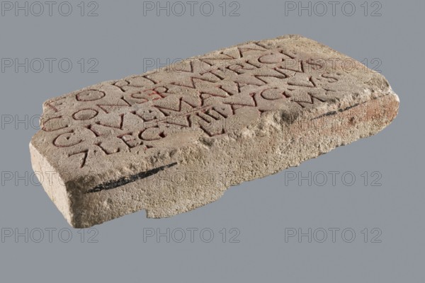 Slab with a Latin votive inscription for Fortuna Conservatrix. Limestone slab with a smoothed front surface and sides, and a rather irregular back; upper left corner missing. Carved inscription in 5 lines on the front; letters were at one point turn of the 19th–20th centuries highlighted with red paint two types: a darker paint in most letters and a lighter, very thick paint in the letter E in the second line. Inscription names Caius Iulius Maianus 2nd–3rd century. Subject: Fortuna mythology; related to baths, centurions, Roman legions, and Germania Superior province. Votive slab, dimensions: width 33 cm, height 18 cm, thickness 6 cm. Date: 130–259/260., A rectangular stone slab with a rough, weathered surface sits against a plain gray background, showing beige and light brown tones with patches of darker brown and subtle reddish areas; the top face has carved grooves filled with a muted red pigment that follow linear patterns, and there is a thin dark streak along one of the slab's lo...