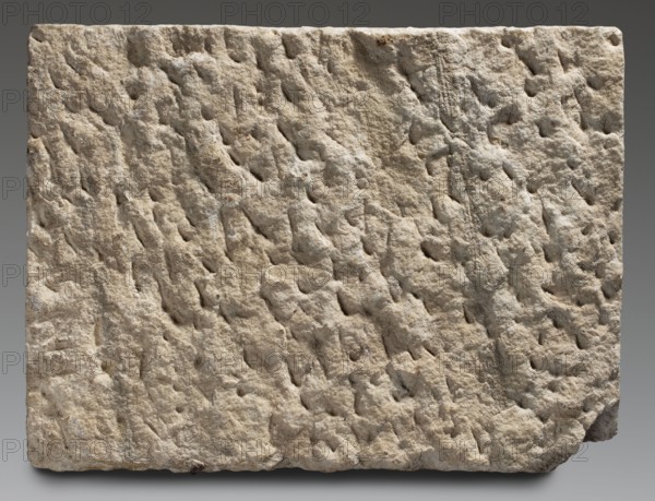 Tombstone of Marcus Julius Sistus with bilingual inscription, unknown Roman workshop; sepulchral funerary architecture, Greek inscriptions, Latin inscriptions, imperial freedmen. Rectangular marble slab with a very irregularly worked back surface. On the front surface are traces of numerous auxiliary lines vertical and horizontal, into which three lines of Latin text and three lines of Greek text are rather unevenly inscribed. Date: after 14 AD–end of 1st century 1st century. Place of origin: Italy, possibly Rome. Slab: height 42 cm, width 56 cm, depth 5 cm, weight 29 kg., A roughly rectangular block with an uneven, pitted surface showing a rough, weathered texture; the block displays a range of beige and tan tones with areas of light gray and small darker brown spots; the edges are irregular with one lower corner chipped, and the background is a smooth gradient of muted gray.