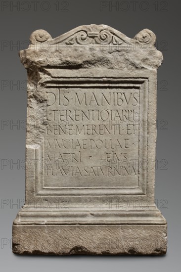Funerary altar of Lucius Terentius Tarpius and Mucia Polla with Latin inscription. Unknown Roman workshop. At the top is a channel for a liquid offering. Sides decorated with representations of sacrificial vessels – a jug and a platter with the head of Medusa. End of the 1st century previously dated to 2nd half of the 1st century–1st half of the 2nd century. Roman Empire 27 BC–476. Sepulchral funerary architecture; Latin inscriptions. Altar; height 90 cm, width 59 cm, depth 44 cm, weight 480 kg., A carved stone slab standing upright with a rectangular central panel bordered by raised molding and set on a stepped base, the top edge shaped with a curved, decorative crest featuring spiral motifs; the stone surface is light gray with areas of darker gray and beige weathering, showing rough, chipped edges and a few pitted spots, while the central panel appears smoother than the surrounding frame; overall the object has an aged, worn texture with subtle shadows emphasizing its carved relief.