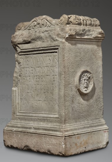 Funerary altar of Lucius Terentius Tarpius and Mucia Polla with Latin inscription, unknown Roman workshop; classified Gallic scenography; Medusa mythology; Latin inscriptions; sepulchral funerary architecture. At the top is a channel for a liquid offering. Sides decorated with representations of sacrificial vessels – a jug and a platter with the head of Medusa. End of the 1st century previously dated 2nd half of the 1st century–1st half of the 2nd century, Roman Empire 27 BC–476. Altar. Height 90 cm, width 59 cm, depth 44 cm, weight 480 kg., A stone rectangular monument with a weathered surface in shades of gray and beige, showing carved decorative scrollwork along the top edge and a raised framed panel on one side with an incised area. The front corner and edges appear chipped and rough, and the base is broader with layered molded profiles. One side features a small circular relief with a carved human face surrounded by stylized hair or foliage. The stone shows variations of light gra...
