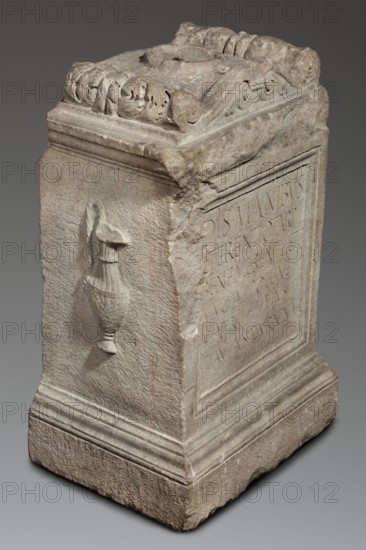 Funerary altar of Lucius Terentius Tarpius and Mucia Polla with Latin inscription, unknown Roman workshop. At the top is a channel for a liquid offering. Sides decorated with representations of sacrificial vessels – a jug and a platter with the head of Medusa. End of the 1st century previous dating: 2nd half of the 1st century–1st half of the 2nd century, Roman Empire 27 BC–476. Type: altar; function: sepulchral funerary architecture., classified Gallic scenography, Medusa mythology, Latin inscriptions. Dimensions: height 90 cm, width 59 cm, depth 44 cm; weight 480 kg., A carved rectangular stone pedestal with weathered surfaces in shades of gray and beige, featuring ornamental scrollwork and rounded decorative forms along the top edge, a raised recessed panel on one side, and a molded stepped base; one visible side shows a relief of a draped vessel or urn projecting from the surface, and the stone displays rough textured areas, chips, and worn corners.