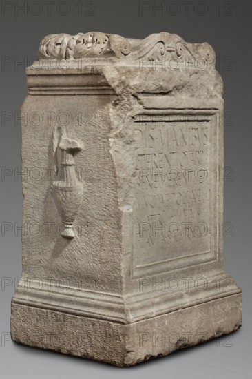 Funerary altar of Lucius Terentius Tarpius and Mucia Polla with Latin inscription, unknown Roman workshop. At the top is a channel for a liquid offering. Sides decorated with representations of sacrificial vessels – a jug and a platter with the head of Medusa. End of the 1st century previously dated 2nd half of the 1st century–1st half of the 2nd century. Roman Empire 27 BC–476. Altar; sepulchral funerary architecture; Latin inscriptions. Height 90 cm, width 59 cm, depth 44 cm, weight 480 kg., A weathered stone pedestal with a rectangular block shape sits against a neutral gray background, showing rough and smooth carved surfaces in shades of gray, beige, and light brown. The top edge has a decorative carved band with scroll and leaf motifs, and the upper corner appears chipped and worn. One visible side features a raised, carved vessel with a draped cloth, rendered in the same stone color but casting subtle shadows. Another face shows a recessed rectangular panel that is smoother than...