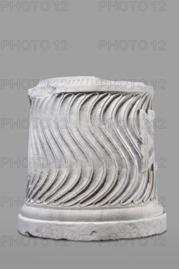 Urn of Gnaeus Pedanius Callistus with Latin inscription, unknown Roman workshop. A cylindrical urn placed on a smooth plinth and a convex base, shaped and decorated to resemble a fragment of a column shaft, tapering towards the top. Entire surface decorated with S-shaped fluting strigillea, closed at top and bottom by a narrow frame. In the central part, slightly closer to the cornice than the base, is a tabula ansata with open handles containing the inscription. Inside, a deep niche for ashes was chiseled out. The opening is topped by a molding decorated with radially arranged grooves; the place where the lid was attached is visible. Lid: missing; analogous example with original conical lid suggests this urn likely had a similar conical or possibly domed lid. Inscription letter height 15 cm. Dating: beginning of the 3rd century previously assigned late 2nd–3rd century AD. Probable findplace: Rome; Roman Empire 27 BC–476. Urn Roman, weight 121.5 kg., A sculpted cylindrical stone object...
