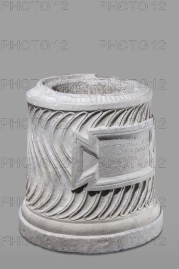 Urn of Gnaeus Pedanius Callistus with Latin inscription, unknown Roman workshop. A cylindrical urn placed on a smooth plinth and a convex base. Its shape and decoration resemble a fragment of a column shaft, tapering towards the top. Entire surface decorated with S-shaped fluting strigillea, closed at top and bottom by a narrow frame. In the central part, slightly closer to the cornice than the base, is a tabula ansata with open handles containing the inscription. Inside the urn a deep niche for ashes was carved out with a chisel. The opening is topped by a molding decorated with radially arranged grooves. The place where the lid was attached is visible. Lid: missing; comparable example with original conical lid suggests this urn likely had a similar conical or possibly domed lid. Inscription letters height 15 cm. Dating: beginning of the 3rd century previously late 2nd–3rd century AD. Probable findspot: Rome, Roman Empire 27 BC–476. Function: funerary urn. Weight 121.5 kg., A cylindri...
