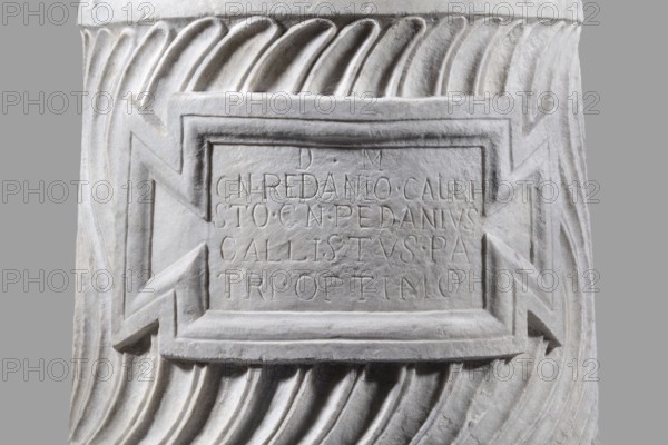 Urn of Gnaeus Pedanius Callistus with Latin inscription, unknown Roman workshop. A cylindrical urn placed on a smooth plinth and a convex base. Its shape and decoration resemble a fragment of a column shaft, tapering towards the top. Entire surface decorated with S-shaped fluting strigillea, closed at top and bottom by a narrow frame. In the central part, slightly closer to the cornice than the base, is a tabula ansata with open handles containing the inscription. Inside the urn a deep niche for ashes was chiseled out. The opening is topped by a molding decorated with radially arranged grooves. The place where the lid was attached is visible. Lid: missing; a comparable urn is known with an original conical lid, so this urn likely had a similar conical or possibly domed lid. Inscription: letter height 15 cm. Dating: beginning of the 3rd century previously dated late 2nd–3rd century AD. Probable findspot: Rome; origin: Roman Empire 27 BC–476. Type: Roman urn. Weight: 121.5 kg., A carved ...