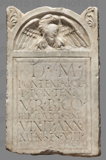 Tombstone, unknown Roman workshop; 2nd half of the 1st–1st half of the 2nd century; Rome findspot; slab, height 34 cm, width 21 cm, depth 5 cm, weight 10 kg., A rectangular carved stone slab with a rounded top featuring a relief of a bird with outstretched wings, flanked by carved leaf-like motifs at the upper corners; the slab is pale beige to off-white with areas of darker beige and light brown weathering, and shows incised rectangular framing and a recessed panel below the bird containing multiple rows of carved depressions; the surface has chisel marks, slight chips and worn edges, and a rougher base with some irregularities.