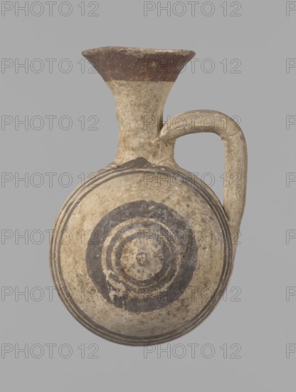 Cypriot gourd jug. A small vessel with a barrel-shaped gourd-shaped body, without a foot, with a spherical bottom. The center is marked with a small protruding point on both short sides. A single handle on the long side, mounted at the greatest bulge of the body and at the base of the neck, bent at a right angle. Quite long neck, strongly flaring, conical spout. Neck and handle slightly offset from the vessel axis; the handle is mounted at a slight angle, with a very clear trace of finger pressure on the right side where it is attached to the neck. Reddish-brown clay. Yellowish-brown engobe. Decoration in black-brown and matte red. The rim is decorated externally with a wide matte red band. The body is decorated on both sides with two groups of three concentric black-brown lines, of varying widths and spacing. In the groups on the shorter sides, the outer line is much wider than the others. Protruding points are marked in black-brown. Preservation condition: cracked and glued neck and ...