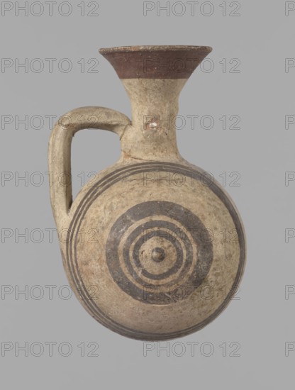 Cypriot gourd jug, A small vessel with a barrel-shaped gourd-shaped body, without a foot, with a spherical bottom. The center is marked with a small protruding point on both short sides. A single handle on the long side, mounted at the greatest bulge of the body and at the base of the neck, bent at a right angle. Quite long neck, strongly flaring, conical spout. Neck and handle slightly offset from the vessel axis; the handle is mounted at a slight angle, with a very clear trace of finger pressure on the right side where it is attached to the neck. Reddish-brown clay. Yellowish-brown engobe. Decoration in black-brown and matte red. The rim is decorated externally with a wide matte red band. The body is decorated on both sides with two groups of three concentric black-brown lines, of varying widths and spacing. In the groups on the shorter sides, the outer line is much wider than the others. Protruding points are marked in black-brown. Preservation condition: cracked and glued neck and ...