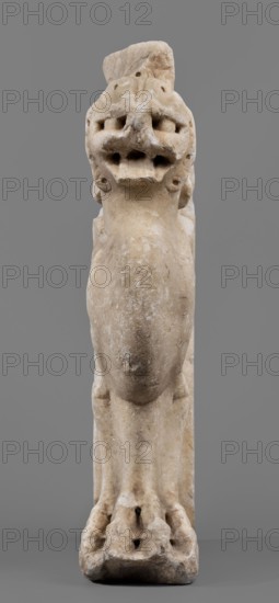Bench support with a griffin figure, unknown Roman workshop; bench support decorated with a winged lion; mid-1st century, Roman Empire 27 BC–476; Roman sculpture; furniture; griffins myth.; table or bench support; height 46 cm, width 32 cm, depth 9 cm, weight 19.7 kg., A pale beige stone sculpture of an animal-like figure is shown against a solid gray background; the figure appears tall and vertical with a rounded elongated central body, a textured surface with weathered and slightly darker beige patches, and a head featuring deep-set hollowed areas and multiple rounded protrusions. The lower portion shows limb-like forms leading to a broad, irregular base with carved indentations and darker spots, and the overall colors visible are various shades of beige, off-white, and gray.