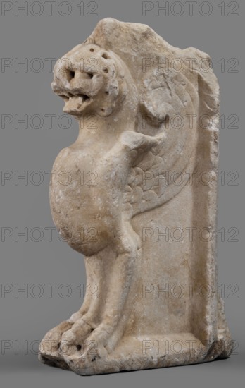 Bench support with a griffin figure, unknown Roman workshop; furniture; griffins myth.; Roman sculpture; table or bench support decorated with a winged lion, ca. mid-1st century, Roman Empire 27 BC–476; furniture, sculpture; height 46 cm, width 32 cm, depth 9 cm, weight 19.7 kg, A carved stone sculpture of a winged creature mounted on a rectangular stone backing, shown against a gray background; the creature has a rounded belly, a lion-like head with an open mouth and visible carved holes on the face, a pair of carved wings with feather-like patterns, forelimbs and hind limbs ending in clawed paws that rest on a flat base, and an overall weathered surface with variations of beige, tan, off-white, and light brown tones.