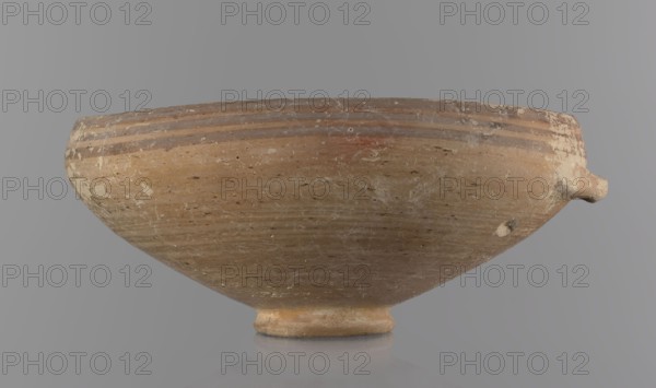 Bowl, excavations at Nea Paphos Cyprus — Bronze Age, 1650–1200 BC. Ceramic footed bowl/goblet on a low ring-shaped stem; walls diverge at a strongly obtuse angle and bend slightly inward at three-quarters height with a small horizontal vise at the bend. Light-sand clay with light-brown exterior paint and brown-red interior. Exterior painted decoration: narrow horizontal lines slightly darker than the ground, two horizontal purple stripes under the rim, a stripe covering the rim edge, and a belly stripe just below the rim. Interior with numerous light purple horizontal lines. Origin: Cyprus. Dimensions: height 8 cm, width 17 cm., A shallow, rounded ceramic bowl resting on a small foot, shown from the side against a neutral gray background, with a short protruding spout on one side; the bowl surface shows a weathered, rough texture with scratches and flaking, and has a base color of light brown and tan with areas of darker brown streaks and mottled white patches, with several horizontal ...