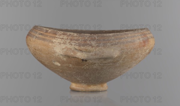 Bowl, excavations at Nea Paphos Cyprus - equivalent. Goblet on a low, ring-shaped stem; walls diverge at a strongly obtuse angle and bend slightly inward at three-quarters height with one small horizontal vise at the bend. Light-sand clay covered with light-brown paint outside and brown-red inside. Painted exterior decoration: narrow horizontal lines slightly darker than the ground, two horizontal purple stripes under the rim, a stripe covering the edge of the rim; belly just below the rim. Inside: numerous light purple horizontal lines. Cypriot pottery, Bronze Age, 1650–1200 BC. Vessel goblet, height 8 cm, width 17 cm., A shallow rounded bowl with a short foot, shown from the front against a neutral gray background, displaying an aged surface with worn patches and areas of flaking; the bowl is primarily tan and light brown with bands of darker brown near the rim, and there are patches of off-white residue and small speckles of darker brown and reddish-brown across its exterior.