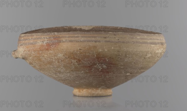 Bowl, excavations at Nea Paphos Cyprus — Bronze Age, 1650–1200 BC. Ceramic footed bowl goblet on a low ring-shaped stem; walls diverge at a strongly obtuse angle and bend slightly inward at three-quarters height with one small horizontal vise at the bend. Light-sand clay with light-brown paint outside and brown-red inside. Painted exterior decoration: narrow horizontal lines slightly darker than ground, two horizontal purple stripes under the rim, a stripe covering the rim edge; belly just below the rim. Interior: numerous light purple horizontal lines. Vessel goblet, height 8 cm, width 17 cm., A shallow, rounded ceramic bowl with a small rounded foot sits against a smooth background; the bowl shows a weathered surface with worn patches and flaking, and a small protruding spout on one side. The exterior displays horizontal banding near the rim and areas of uneven coloration in shades of tan, beige, brown, reddish-brown, and pale off-white. The interior rim appears darker with muted bro...
