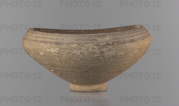 Bowl, excavations at Nea Paphos Cyprus; Bronze Age, 1650–1200 BC. Goblet on a low, ring-shaped stem; walls diverge at a strongly obtuse angle, bending slightly inward at three-quarters height with one small horizontal vise at the bend. Light-sand clay with light-brown paint outside and brown-red inside. Exterior painted decoration: narrow horizontal lines slightly darker than the ground, two horizontal purple stripes under the rim, a stripe covering the rim edge, and a belly stripe just below the rim. Interior has numerous light purple horizontal lines. Vessel type: footed bowl/goblet. Dimensions: height 8 cm, width 17 cm. Origin: Cyprus., A shallow rounded bowl with a small circular foot sits against a smooth gray background, the bowl showing a weathered surface with patches of lighter and darker areas; its overall color palette includes beige, tan, brown, cream, and hints of gray, with several horizontal bands near the rim and a worn, textured exterior.