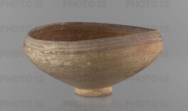 Bowl, excavations at Nea Paphos Cyprus; Bronze Age 1650–1200 BC. Goblet on a low, ring-shaped stem; walls diverge at a strongly obtuse angle and bend slightly inward at three-quarters height with a small horizontal vise at the bend. Light-sand clay with light-brown paint outside and brown-red inside. Painted decoration outside: narrow horizontal lines slightly darker than the ground, two horizontal purple stripes under the rim, a stripe covering the rim edge, and a belly decoration just below the rim. Inside: numerous light purple horizontal lines. Vessel goblet, height 8 cm, width 17 cm., A shallow, rounded ceramic bowl with a narrow circular foot sits against a smooth gray background, showing worn and flaked surfaces. The exterior and interior of the bowl display earthy tones including beige, tan, brown, and muted reddish-brown, with areas of lighter cream-colored flaking. The rim is slightly irregular and the body has subtle horizontal ridges and weathered patches that reveal variat...