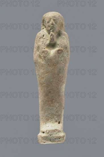 Ushabti, mummy-shaped figurine, funerary gift, hoes. Mummy-shaped figurine standing on a base, back supported by a narrow flat pillar. Arms crossed on the chest, right arm folded over the left; both hands hold hoes. Smooth tripartite wig, round face, facial details illegible, long straight beard. Not inscribed. Egypt; ushabti; height 11.8 cm, width 3.3 cm., A single elongated statue-like object stands against a plain gray background, with a rounded top tapering down to a flared base; the surface appears rough and pitted with a weathered texture and irregular small holes. The object shows a range of muted colors including beige, light brown, pale green, and hints of gray, with darker brown spots and lighter creamy areas. Near the upper portion there are shallow, worn contours suggesting a simplified facial area and crossed arms, with subtle raised and recessed areas around the midsection. The overall shape is narrow and slightly tapered, and the base is broader and slightly squared with...