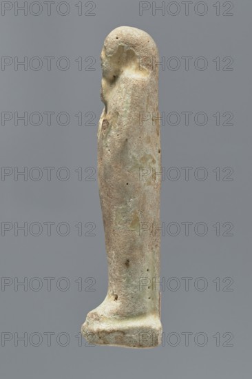 Ushabti, mummy-shaped figurine, funerary gift; standing on a base, back supported by a narrow flat pillar. Arms crossed on the chest, right folded over left; both hands hold hoes. Smooth tripartite wig, round face, facial details illegible, long straight beard. Not inscribed. Egypt; height 11.8 cm, width 3.3 cm., A slender upright figurine viewed in profile against a muted gray background, with a rounded head and a long tapered body standing on a flat rectangular base; the surface shows a rough, pitted texture with irregular patches of pale beige, light tan, and a few areas of muted green, along with some darker brownish spots, and the contours suggest a simple, stylized form with minimal detailing.
