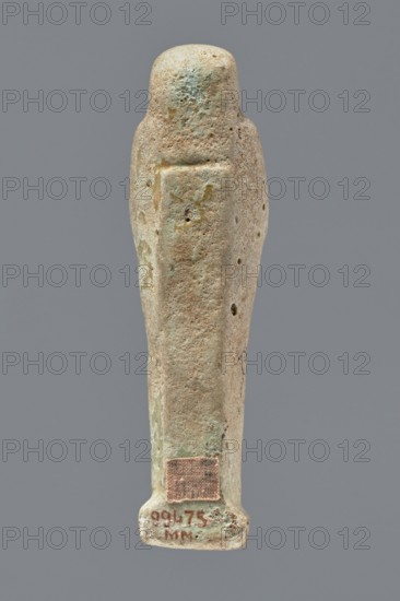 Ushabti, mummy-shaped figurine, funerary gift, hoes. Mummy-shaped figurine standing on a base, the back supported by a narrow, flat pillar. Arms crossed on the chest, the right folded over the left; both hands hold hoes. Smooth tripartite wig, round face with illegible facial details, long straight beard. Not inscribed. Unknown creator; Egypt; ushabti; height 11.8 cm, width 3.3 cm., A vertically oriented carved figure resembling a stylized human form with an elongated body and a rounded head, shown from the back; the surface appears pitted and weathered with a rough, porous texture and small holes; the object displays muted colors including beige, light tan, pale green, and hints of bluish-green patina, with a small darker rectangular patch near the lower end; the background is a uniform medium gray.