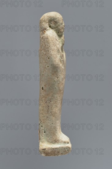 Ushabti, mummy-shaped figurine, funerary gift; standing on a base with the back supported by a narrow flat pillar. Arms crossed on the chest, right folded over left; both hands hold hoes. Smooth tripartite wig, round face, facial details illegible, long straight beard. Not inscribed. Egypt; ushabti; height 11.8 cm, width 3.3 cm., A slender upright carved figure rendered in a pale beige to light tan stone with patches of muted green and darker brown speckling, showing a rounded head, simplified facial profile, and an elongated body that tapers into a flat rectangular base; the surface is pitted with a few small holes and weathered texture, and the background is a uniform mid-gray.