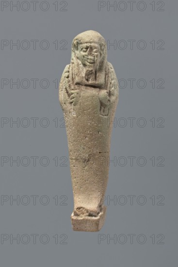 Ushabti, Mummy-shaped figurine, standing on a base with the back supported by a flat pillar. The arms are crossed on the chest and placed at the same level, some distance apart. In each hand are hoes; the right hand also holds a string from which a basket is slung over the left shoulder. The tools are engraved. On the head is a smooth, three-part wig. The face is elongated, with clearly defined eyes, a long nose, and a slight smile. A long, straight beard. Not inscribed. 525–343 BC; Late Period, 27th–30th Dynasty, Egypt. Ushabti; height 9.6 cm., A small sculpted figure with a rounded head and a rudimentary facial carving, arms crossed over the chest, and a tapered lower body ending on a rectangular base; the surface appears textured and pitted with visible cracks and chips, and the predominant colors are beige and light brown with hints of pale green against a solid gray background.