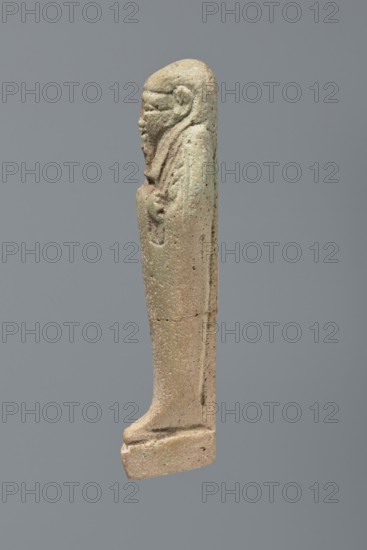 Ushabti, Mummy-shaped figurine, standing on a base with the back supported by a flat pillar. Arms crossed on the chest, placed at the same level and some distance apart; each hand holds hoes. A string in the right hand suspends a basket over the left shoulder. Tools are engraved. Head wears a smooth, three-part wig. Elongated face with clearly defined eyes, a long nose, and a slight smile; long straight beard. Not inscribed. 525–343 BC, Late Period, 27th–30th Dynasty, Egypt. Ushabti; height 9.6 cm., A small upright figurine carved from a light tan to beige material with areas of pale green patina, shown in profile against a plain gray background; the figure has a rounded head, a defined facial profile with a prominent nose and chin, a textured beard, and a long cloak draped down the body, with the hands positioned at the front; the surface shows pitting and small holes, a horizontal crack around the midsection, and slightly darker spots and shadows that give subtle variations in the ta...