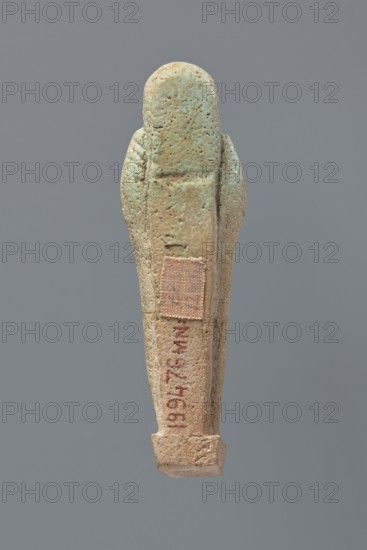 Ushabti, Mummy-shaped figurine, standing on a base, the back supported by a flat pillar. The arms are crossed on the chest and placed at the same level, some distance apart. In each hand are hoes; in the right hand is also a string from which a basket is slung over the left shoulder. The tools are engraved. On the head is a smooth, three-part wig. The face is elongated with clearly defined eyes, a long nose, and a slight smile; a long, straight beard. Not inscribed. 525–343 BC; Late Period, 27th–30th Dynasty, Egypt. Ushabti; height 9.6 cm., An elongated rounded object resembling a small carved figure stands against a plain gray background, showing a weathered surface with mottled green and beige tones and areas of light brown; the surface appears pitted and textured with a rectangular patch of fabric attached to one side and a narrow groove running vertically, with the lower end forming a slightly wider squared base.