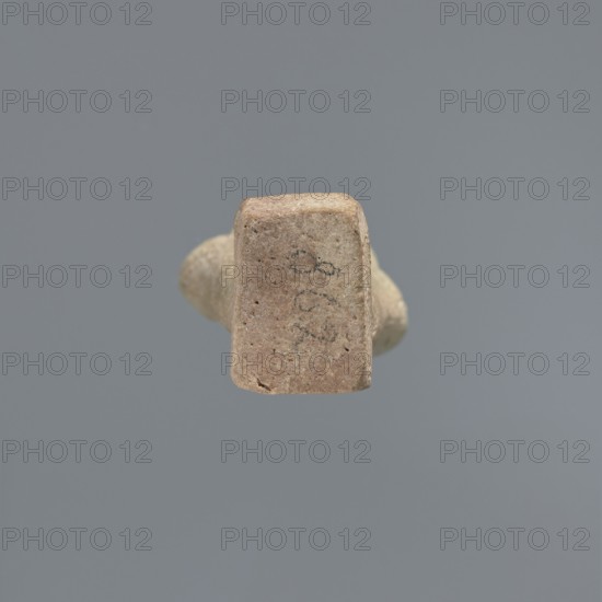 Ushabti, Mummy-shaped figurine, standing on a base, the back supported by a flat pillar. The arms are crossed on the chest and placed at the same level, some distance apart. In each hand are hoes; in the right hand is also a string from which a basket is slung over the left shoulder. The tools are engraved. On the head is a smooth, three-part wig. The face is elongated, with clearly defined eyes, a long nose, and a slight smile on the mouth. A long, straight beard. Not inscribed. 525–343 BC; Late Period, 27th–30th Dynasty, Egypt. Ushabti, height 9.6 cm., A small, irregularly shaped object with a rough, sandy surface in tan and light brown tones is centered against a uniform gray background; the object shows a rectangular face facing forward with some darker speckles and faint dark markings, and a rounded, tapering body extending behind it in similar tan and beige colors.