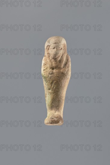 Shabti, Mummy-shaped figurine shabti, funerary gift, made 525–343 BC Late Period, 27th–30th Dynasty, Egypt. Standing on a base, back supported by a flat pillar. Arms crossed on the chest, holding hoes in both hands; a string in the right hand suspends a basket slung over the left shoulder. Tools slightly convex. Wearing a smooth, tripartite wig; round face with clearly defined large eyes; mouth and nose not visible. Not inscribed. Height 7 cm., A single small statuette is centered against a plain gray background, shaped like a tapered figure with a rounded head and simplified facial features; the surface appears textured and worn with a weathered, uneven finish. The object shows a mix of beige and brown tones overall, with areas of muted greenish discoloration and darker brown patches; the head area is slightly lighter beige while the lower portion has warmer brown shading. The form narrows toward the base and flares slightly at the bottom, and the surface displays irregularities and s...