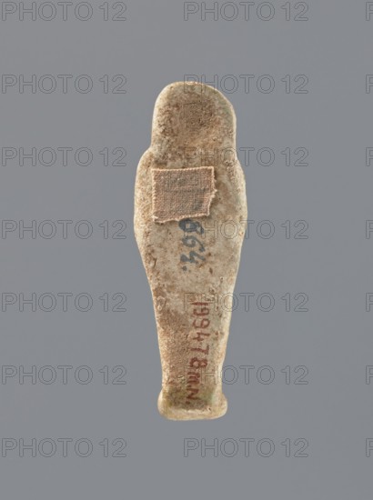 Shabti, mummy-shaped figurine, funerary gift, hoes; standing on a base with back supported by a flat pillar. Arms crossed on the chest, holding hoes in both hands; in the right hand a string suspends a basket over the left shoulder. Slightly convex tools. Smooth, tripartite wig, round face with clearly defined large eyes; mouth and nose not visible. Not inscribed. 525–343 BC; Late Period, 27th–30th Dynasty, Egypt. Shabti, height 7 cm., A roughly human-shaped small object with a rounded top and tapered body in beige and light brown tones, showing a textured, worn surface and patches of darker brown and greenish discoloration, with a small square of coarse fabric attached near the upper middle in a tan color, all set against a uniform gray background.