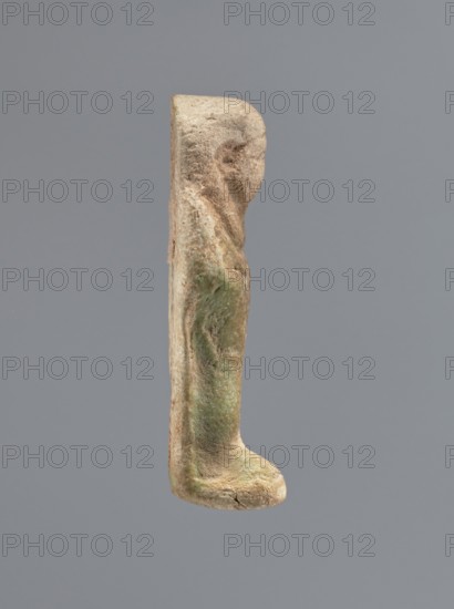 Shabti, mummy-shaped figurine shabti, funerary gift, standing on a base, back supported by a flat pillar. Arms crossed on the chest, holding hoes in both hands; a string in the right hand suspends a basket over the left shoulder. Slightly convex tools. Wears a smooth tripartite wig. Round face with clearly defined large eyes; mouth and nose not visible. Not inscribed. 525–343 BC; Late Period, 27th–30th Dynasty, Egypt. Height 7 cm., A narrow vertical object with a textured, weathered surface shown against a plain gray background; the object is predominantly beige and light brown with areas of greenish patina and darker brown patches, featuring an irregular rounded top, a slightly contoured midsection, and a thicker rounded base with a small crack.