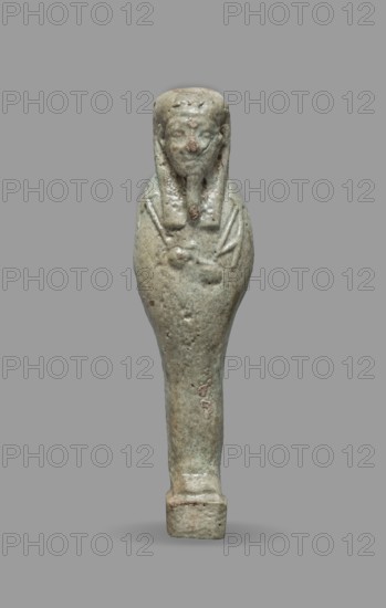 Ushabti, Mummy-shaped funerary figurine standing on a base, back supported by a flat pillar. Arms crossed on the chest, hands visible from under the shroud with the right slightly above the left; the left hand holds a pickaxe, the right a hoe and a string from which a basket is slung over the left shoulder. Convex tools. Smooth three-part wig, triangular face, clearly marked large eyes, wide nose, mouth in a slight grimace. Not inscribed. Late Period, 664–332 BC, Egypt. Shabti; height 9.3 cm, width 2.5 cm., A small sculpted figure shown against a plain gray background, depicting a mummiform person with a rounded head and a stylized beard, the surface appearing weathered and pitted with rough patches; the figure is predominantly a muted greenish-gray with areas of brownish discoloration and lighter pale spots, the head has long flanking elements over the shoulders and faint incised lines suggesting folded arms across the chest, the lower body tapers into a narrow leg-like form that ends...