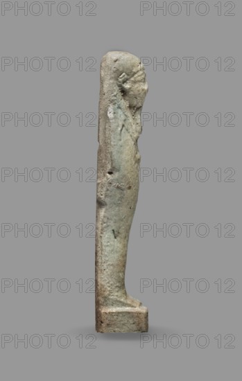 Ushabti, Mummy-shaped figurine standing on a base, back supported by a flat pillar. Arms crossed on the chest, hands visible from under the shroud, right slightly above the left. Left hand holds a pickaxe, right hand holds a hoe and a string from which a basket is slung over the left shoulder. Convex tools. Smooth, three-part wig; triangular face with clearly marked large eyes, wide nose, mouth in a slight grimace. Not inscribed. Late Period, Egypt, 664–332 BC. Shabti; height 9.3 cm, width 2.5 cm., A narrow vertical stone or ceramic figure shown in profile on a plain gray background, with a rounded head, elongated torso, and a rectangular base; the surface appears weathered and pitted with rough texture, showing a blend of muted gray, greenish, and beige tones, with some darker and lighter speckling and areas of subtle discoloration.