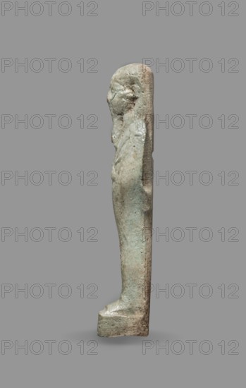 Ushabti, Back pillars, wigs, funerary figurines, hoes, grave goods, mummy-shaped figurines, standing on a base, the back supported by a flat pillar. Arms crossed on the chest. Hands visible from under the shroud, the right slightly above the left. In the left hand a pickaxe, in the right a hoe. In the right hand also a string, on which a basket is slung over the left shoulder. Convex tools. On the head is a smooth, three-part wig. Triangular face, clearly marked large eyes, a wide nose, mouth in a slight grimace. Not inscribed. 664–332 BC; Late Period, Egypt. Shabti; height 9.3 cm, width 2.5 cm., A narrow vertical statuette of a standing human figure shown in profile against a plain background, with a smooth elongated shape and simplified features including a defined head, shoulders, and torso; the surface appears textured and worn with areas of roughness and small pitting, and the form narrows toward the feet which rest on a small rectangular base; visible colors include muted green, ...