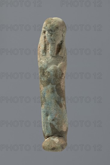 Ushabti, Mummy-shaped figurine, very schematically shaped. Hands crossed on the chest. On the head is a long, tripartite wig. Facial details illegible. Not inscribed. ca. 1069–656 BC; Third Intermediate Period, Egypt. Ushabti; height 9.4 cm., A narrow vertical object resembling a carved figure is centered against a plain gray background; the surface is rough and weathered with a textured, pitted appearance and mottled colors of green, blue-green, tan, beige, and hints of brown; the top area suggests a simplified head shape with shallow facial contours and elongated side forms, while the elongated body beneath shows uneven surface irregularities and discoloration, ending in a rounded base with a visible crack.