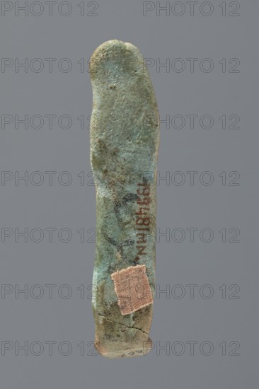 Ushabti, Mummy-shaped figurine, very schematically shaped. Hands crossed on the chest. On the head is a long, tripartite wig. Facial details illegible. Not inscribed. ca. 1069–ca. 656 BC; Third Intermediate Period, Egypt. Ushabti; height 9.4 cm., A single elongated, irregularly shaped object photographed against a plain gray background, with a rough, pitted surface. The object shows a predominant greenish-blue patina with areas of darker green and brown discoloration. Near the lower portion there is a small square patch of tan fabric affixed to the surface. The top end is rounded and the bottom end is slightly wider and shows a visible crack. The surface texture varies from smoother worn areas to more granular, corroded spots.