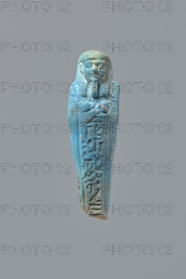 Ushabti, Mummy-shaped figurine, back supported on a flat pillar. Feet missing. Arms crossed on the chest, right folded over left. Hoes in both hands; in the right hand a string on which a basket is slung over the left shoulder. Slightly convex tools. On the head, a smooth, three-part wig. Round face, clearly marked large eyes, wide nose, mouth in a slight grimace. Engraved inscription in one column on the front of the legs text illegible. 404–343 BC; Late Period, 28th–30th Dynasty, Egypt. Ushabti; height 7 cm., A small carved figure shaped like a standing wrapped mummy with crossed arms and a stylized beard and wig, shown against a plain gray background; the object is predominantly turquoise blue with areas of lighter blue and pale green patina and small spots of brownish discoloration and wear.